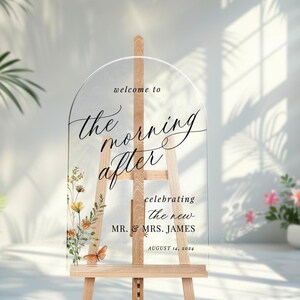 May include: Clear acrylic wedding sign with an arched top, displaying the text "welcome to the morning after" in elegant script. Floral and butterfly accents adorn the lower left. The sign is on a wooden easel, with the date August 14, 2024.