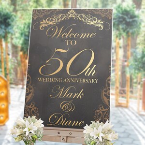 May include: A rectangular black sign with gold text and ornate scrollwork borders. The text reads Welcome TO 50th WEDDING ANNIVERSARY Mark & Diane. The sign is displayed on a wooden easel and decorated with white lilies and baby's breath flowers.