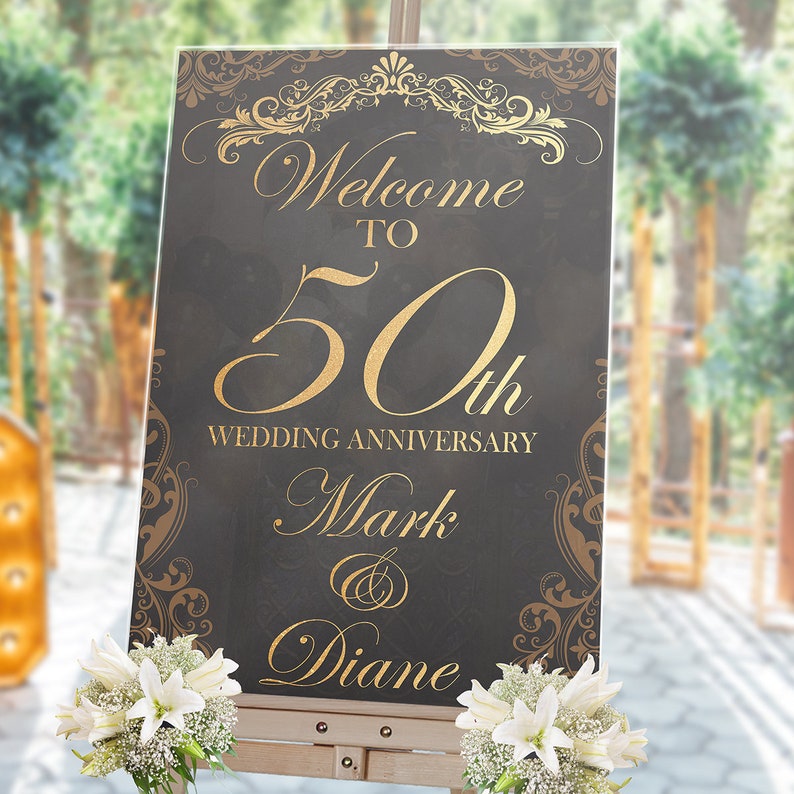 May include: A rectangular black sign with gold text and ornate scrollwork borders. The text reads Welcome TO 50th WEDDING ANNIVERSARY Mark & Diane. The sign is displayed on a wooden easel and decorated with white lilies and baby's breath flowers.