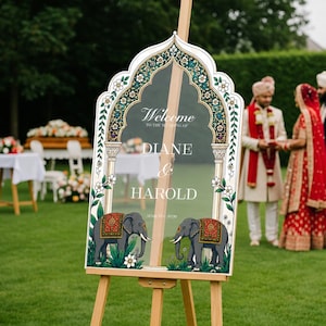 Peut inclure: Un panneau de bienvenue de mariage en acrylique transparent avec un cadre en arche orn&eacute; de motifs floraux verts, dor&eacute;s et blancs. Le texte indique Welcome to the wedding of Diane & Harold. Deux &eacute;l&eacute;phants gris avec des couvertures rouges et dor&eacute;es.