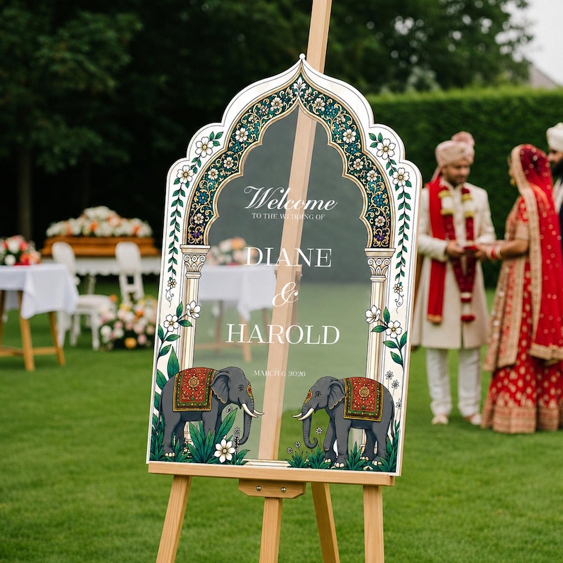 Peut inclure: Un panneau de bienvenue de mariage en acrylique transparent avec un cadre en arche orn&eacute; de motifs floraux verts, dor&eacute;s et blancs. Le texte indique Welcome to the wedding of Diane & Harold. Deux &eacute;l&eacute;phants gris avec des couvertures rouges et dor&eacute;es.