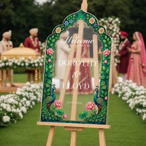 Op de afbeelding: Een transparant acryl welkomstbord voor een bruiloft op een houten ezel met een decoratieve rand van groene bladeren, roze lotusbloemen en twee pauwen. De tekst luidt: Welcome to the wedding of Dorothy & Lloyd, June 6 2026.