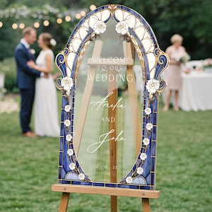 May include: A decorative wedding sign with a blue and gold stained glass-style frame, featuring white floral accents. The sign reads "WELCOME TO OUR WEDDING" and the names "Amelia and John" with the date "10 15 25". The sign is displayed on a wooden easel.
