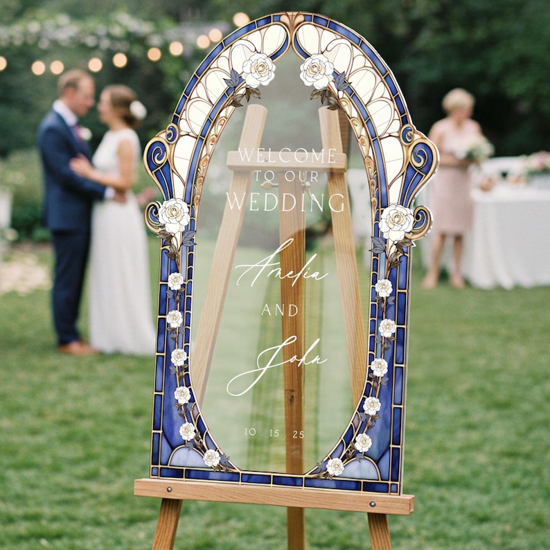 May include: A decorative wedding sign with a blue and gold stained glass-style frame, featuring white floral accents. The sign reads "WELCOME TO OUR WEDDING" and the names "Amelia and John" with the date "10 15 25". The sign is displayed on a wooden easel.