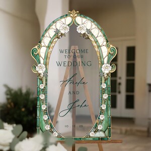 May include: An arched wedding welcome sign featuring a stained glass-style frame in shades of teal and gold with white rose illustrations. The clear centre displays the text: WELCOME TO OUR WEDDING, Amelia AND John, 10.15.25, mounted on a wooden easel.