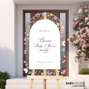May include: A white arched welcome sign on a wooden easel featuring a floral border with colorful painted flowers. The text reads: WELCOME TO Becca's Baby Shower party OCTOBER 22, 2024. The sign is positioned in front of a dark brown door.