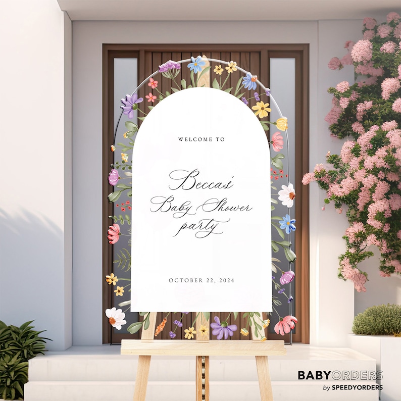 May include: A white arched welcome sign on a wooden easel featuring a floral border with colorful painted flowers. The text reads: WELCOME TO Becca's Baby Shower party OCTOBER 22, 2024. The sign is positioned in front of a dark brown door.