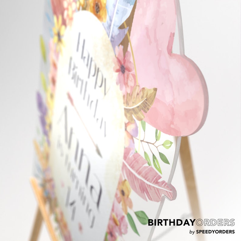 May include: A custom acrylic birthday sign featuring a heart-shaped cutout on the right side. The sign displays watercolor floral illustrations in pink, yellow, and blue, with the text Happy Birthday Anna is turning 14 printed in the center.