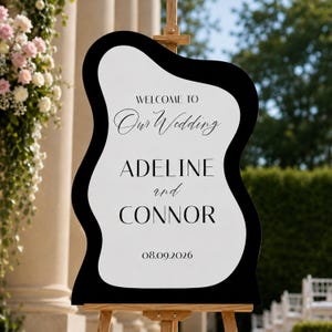 May include: A wavy-edged wedding welcome sign featuring a white center with black borders. The text reads: WELCOME TO Our Wedding ADELINE and CONNOR 08.09.2026. The sign is mounted on a wooden easel, displaying names and a date in black typography.