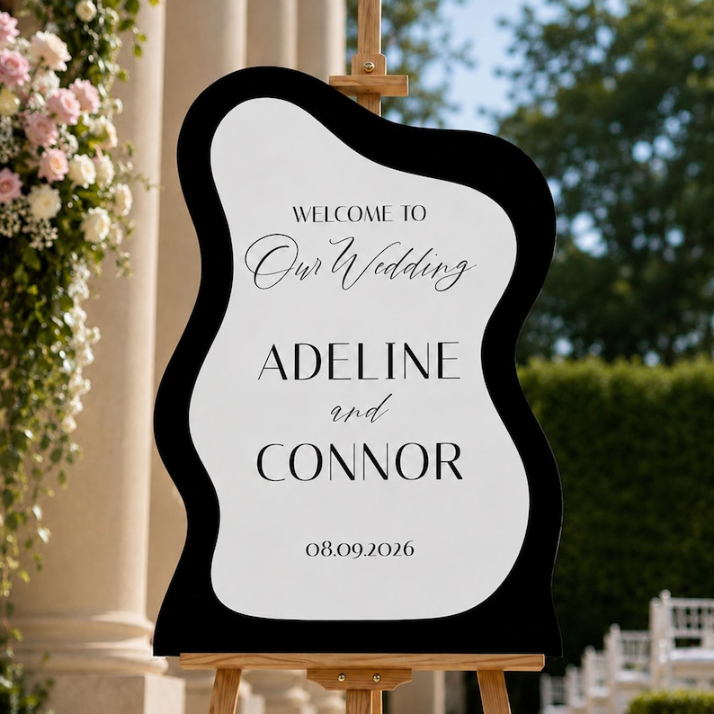 May include: A wavy-edged wedding welcome sign featuring a white center with black borders. The text reads: WELCOME TO Our Wedding ADELINE and CONNOR 08.09.2026. The sign is mounted on a wooden easel, displaying names and a date in black typography.