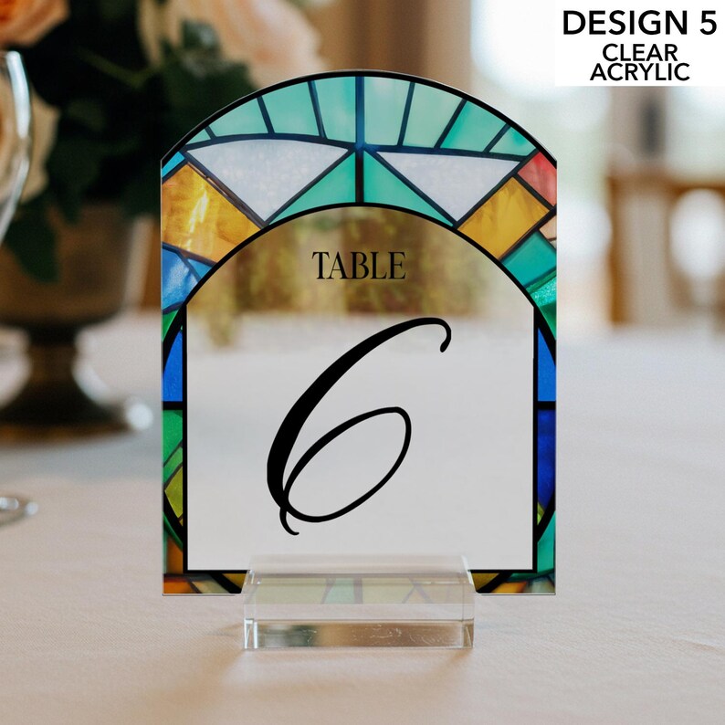 May include: A clear acrylic table number with a stained glass design. The arched top features a colorful pattern of blue, green, yellow, and white. The number 6 is displayed in black, with the word "TABLE" above it. The design is labeled "DESIGN 5 CLEAR ACRYLIC."