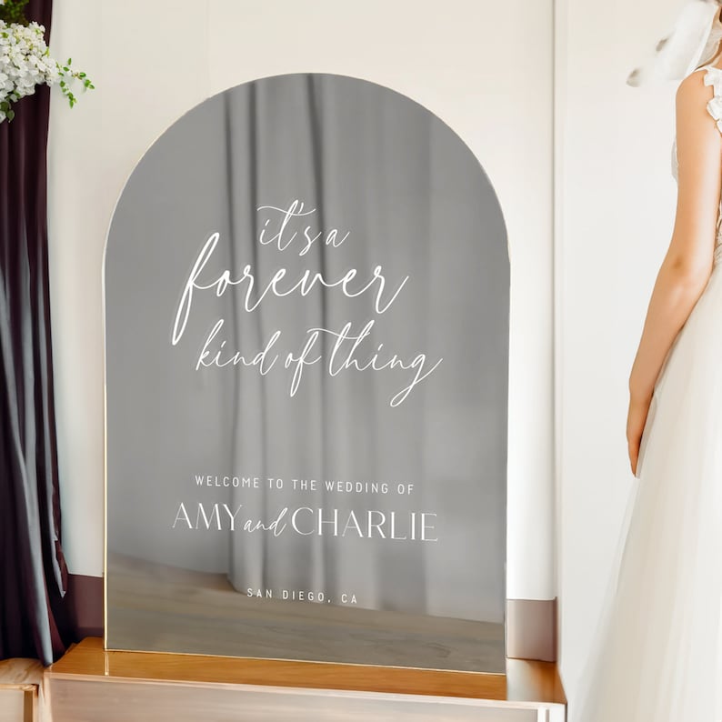 Może przedstawiać: Łukowaty, odblaskowy znak lustrzany z białym napisem: it's a forever kind of thing, WELCOME TO THE WEDDING OF AMY and CHARLIE oraz SAN DIEGO, CA. Znak ma gładką, metaliczną powierzchnię i zaokrągloną g&oacute;rną krawędź.
