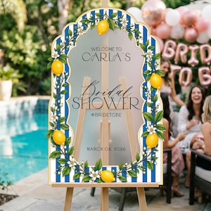 May include: An arched sign with a blue and white striped border, decorated with yellow lemons and white flowers. The text reads: WELCOME TO CARLA'S Bridal SHOWER #BRIDETOBE MARCH 04, 2026. The sign is mounted on a light-colored wooden easel stand.