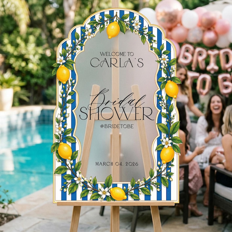 May include: An arched sign with a blue and white striped border, decorated with yellow lemons and white flowers. The text reads: WELCOME TO CARLA'S Bridal SHOWER #BRIDETOBE MARCH 04, 2026. The sign is mounted on a light-colored wooden easel stand.