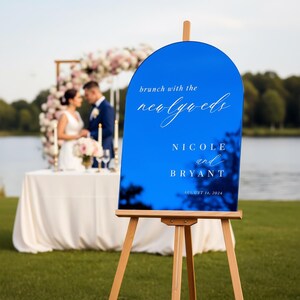 May include: A bright blue arched acrylic wedding sign on a wooden easel. The sign features white text that reads: brunch with the newlyweds, NICOLE and BRYANT, AUGUST 14, 2024. The surface is reflective, showing a blurred outdoor landscape.
