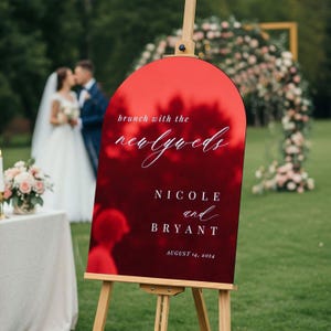 May include: An arched, glossy red sign on a wooden easel featuring white text that reads: brunch with the newlyweds, NICOLE and BRYANT, AUGUST 14, 2024. The sign has a reflective surface showing blurred outdoor scenery and wedding details.