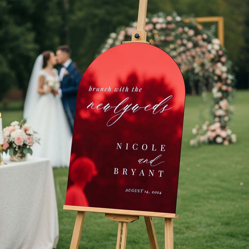 May include: An arched, glossy red sign on a wooden easel featuring white text that reads: brunch with the newlyweds, NICOLE and BRYANT, AUGUST 14, 2024. The sign has a reflective surface showing blurred outdoor scenery and wedding details.
