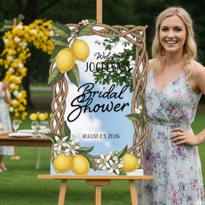 May include: A wooden easel supports a sign with a decorative border of woven branches and lemon illustrations. The sign reads "Welcome to Jocelyn's Bridal Shower" with the date August 03, 2026. A woman in a floral dress smiles.
