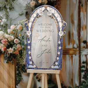 May include: A wooden wedding welcome sign with a stained glass design. The sign features the text "Welcome to our Wedding" above the names "Amelia and John" and the date "10 15 25". The sign is decorated with white roses and blue accents.