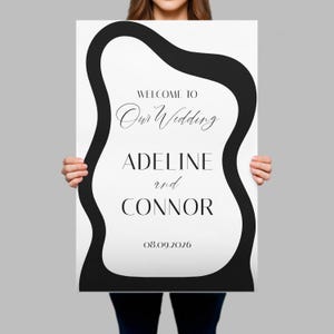 May include: A rectangular wedding sign featuring a white background with a thick, black, wavy border. The text reads: WELCOME TO Our Wedding ADELINE and CONNOR 08.09.2026. The design uses a combination of serif and script fonts in black ink.