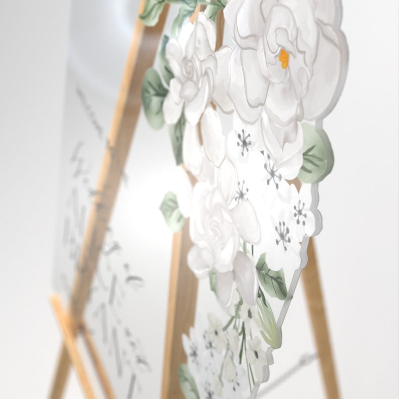 May include: A clear acrylic sign featuring a printed floral design of white gardenias with green leaves and small white blossoms. The sign is mounted on a light-colored wooden easel. The surface displays faint, illegible cursive black text.