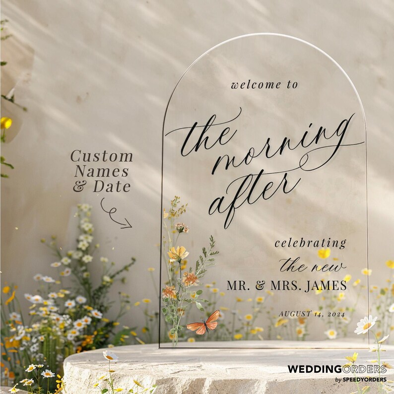 May include: Clear acrylic wedding sign with an arched top. The sign reads "welcome to the morning after" and includes the names "MR. & MRS. JAMES" and the date "AUGUST 14, 2024". Floral and butterfly accents decorate the sign.