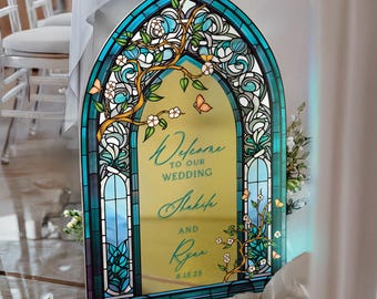 May include: An arched wedding welcome sign featuring a gold mirror center with blue stained glass borders. The design includes white floral branches and orange butterflies. Text reads: Welcome to our wedding, Shakifa and Ryan, 6.15.25.