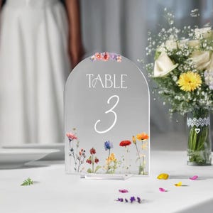 May include: A clear acrylic table number with a floral design and the text "TABLE 3" in white lettering.