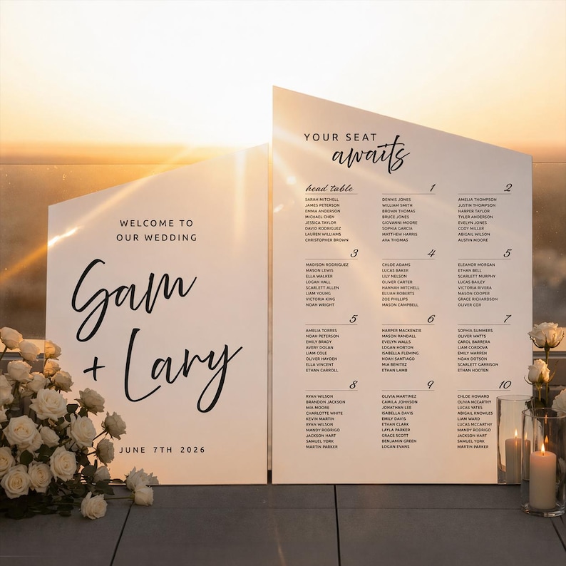 K&ouml;nnte beinhalten: Zwei wei&szlig;e Hochzeitsschilder mit schwarzem Text. Auf einem Schild steht "Willkommen zu unserer Hochzeit" und "Gam + Lary" mit dem Datum "7. Juni 2026". Das andere Schild listet die Namen der G&auml;ste unter "Ihr Platz erwartet Sie" auf. Wei&szlig;e Rosen und Kerzen im Vordergrund.