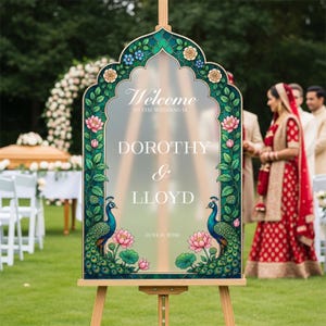Op de afbeelding: Een mat acryl welkomstbord voor een bruiloft met een boogvormige bovenkant, voorzien van een decoratieve rand van groene wijnstokken, roze lotusbloemen en twee blauwe pauwen. De tekst luidt: Welcome TO THE WEDDING OF DOROTHY & LLOYD JUNE 6, 2026.