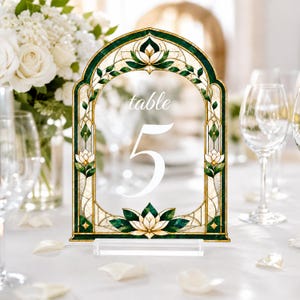 May include: An arched table number sign featuring a stained glass design with deep green, cream, and gold tones. The sign displays the word table in a script font above the large white numeral 5, all supported by a clear rectangular acrylic base.