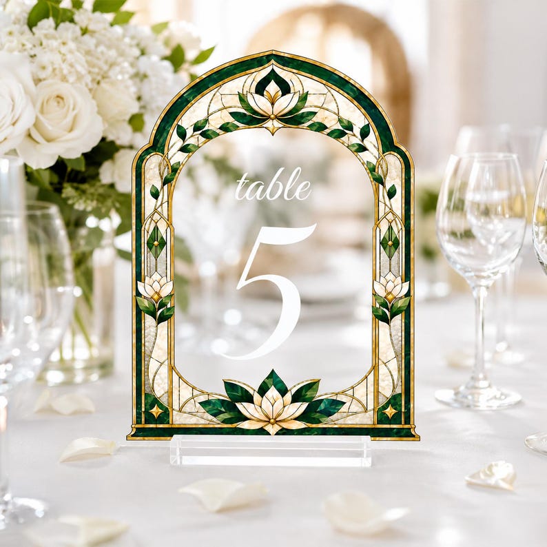 May include: An arched table number sign featuring a stained glass design with deep green, cream, and gold tones. The sign displays the word table in a script font above the large white numeral 5, all supported by a clear rectangular acrylic base.