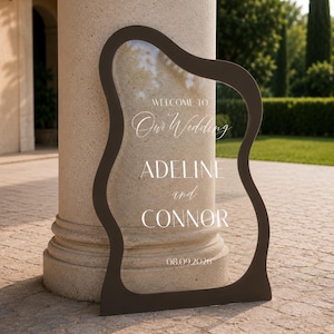 May include: A clear acrylic wedding sign with a dark brown, wavy-edged frame. The text reads: WELCOME TO Our Wedding ADELINE and CONNOR 08.09.2026. The sign is positioned upright, displaying white lettering against the transparent surface.