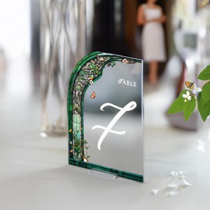 May include: A green and white stained glass design table number with a mirrored surface. The number seven is written in white script on the mirror. The text "Table" is printed in black at the top of the mirror.