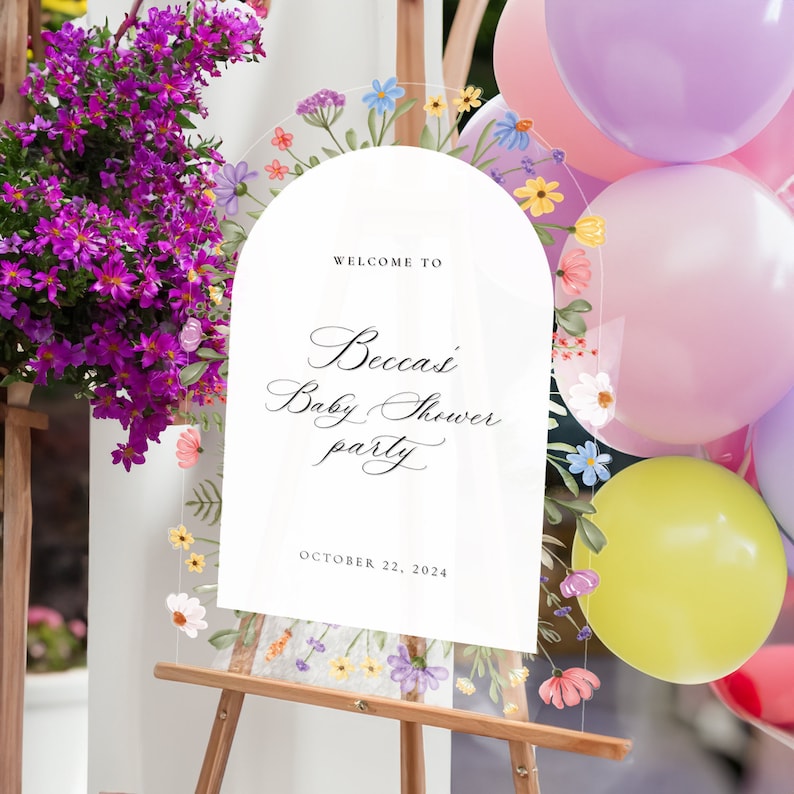 May include: An arched clear acrylic sign featuring a floral border with colorful painted flowers and green leaves. The text reads: WELCOME TO Becca's Baby Shower party OCTOBER 22, 2024. The sign is displayed on a wooden easel with balloons nearby.