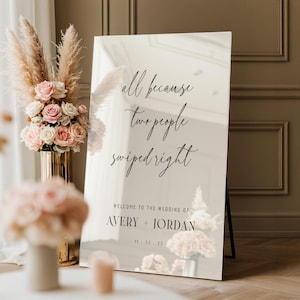 May include: A mirrored wedding sign with the text "all because two people swiped right." The sign also includes the names "Avery + Jordan" and the date "11.22.23". The sign is propped up on a black stand.