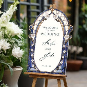 May include: A decorative wedding welcome sign with a stained-glass design. The sign features a blue and gold border with white roses and the text "Welcome to our Wedding" above the names Amelia and John, dated 10.15.25. The sign is displayed on a wooden easel.