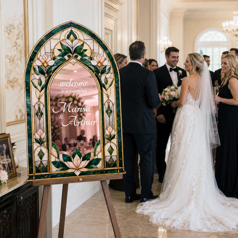 May include: An arched wedding welcome sign featuring a stained glass-style border in shades of dark green and cream with floral motifs. The center mirror displays the text: welcome to our wedding, Marisa & Arthur, 05-02-26, set on a wooden easel.