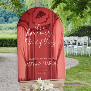 Może przedstawiać: Łukowaty znak powitalny na ślub w odcieniach czerwieni z napisem "it's a forever kind of thing", "WELCOME TO THE WEDDING OF AMY and CHARLIE" oraz "SAN DIEGO, CA". Znak jest umieszczony na drewnianej sztaludze z białymi liliami u podstawy.