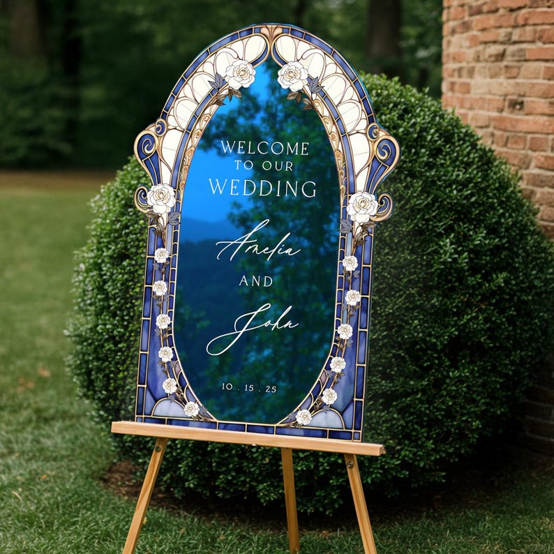 May include: A decorative wedding welcome sign with a blue mirrored surface and an ornate, stained-glass style frame. The sign reads "Welcome to our Wedding" and features the names "Amelia and John" along with the date 10.15.25. It is displayed on a wooden easel.