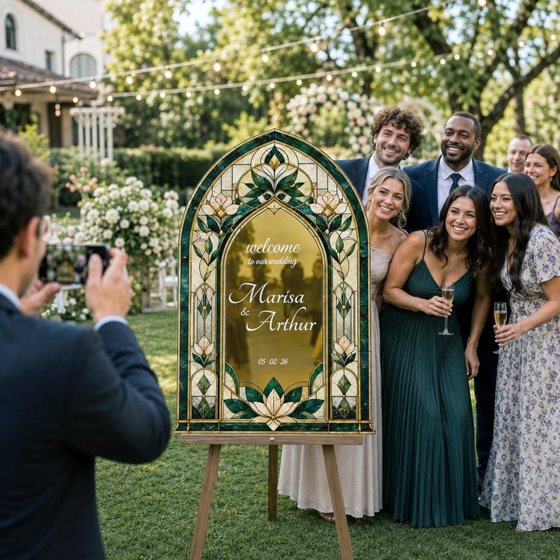 May include: An arched wedding welcome sign featuring a stained glass-style border with green and cream floral patterns. The center is gold with white text reading: welcome to our wedding, Marisa & Arthur, 05.02.26. The sign stands on a wooden easel.