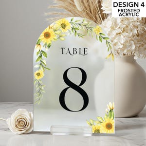 May include: Frosted acrylic table number with a floral sunflower design. The number 8 is prominently displayed in black, with the word "TABLE" above it. The design includes yellow sunflowers and green leaves. The table number is on a clear acrylic base.