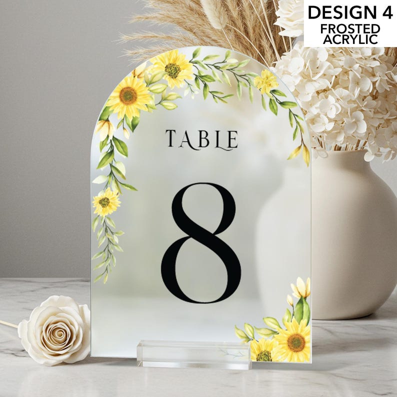 May include: Frosted acrylic table number with a floral sunflower design. The number 8 is prominently displayed in black, with the word "TABLE" above it. The design includes yellow sunflowers and green leaves. The table number is on a clear acrylic base.