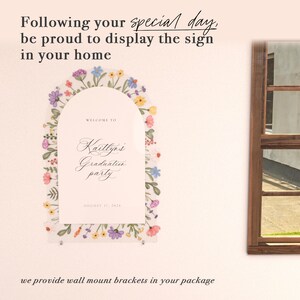 May include: An arched acrylic sign featuring a colorful floral border with purple, blue, yellow, and pink flowers. The text reads: Welcome to Kaitlyn's Graduation party, August 17, 2024. The sign is mounted with silver hardware on a light wall.