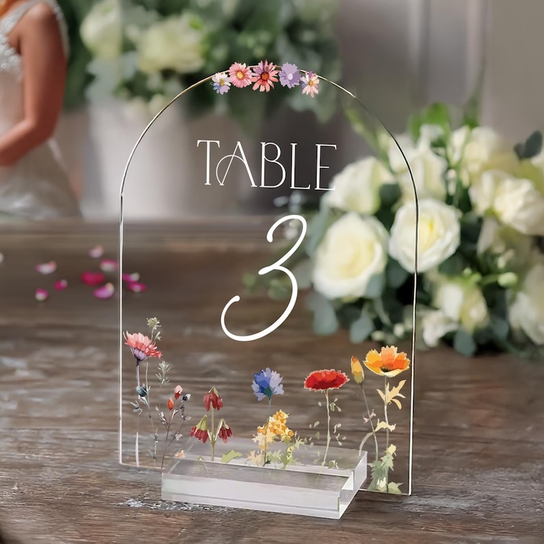 May include: Clear acrylic table number with a floral design. The number 3 is written in white script. The top of the sign has a row of pink, purple, and white flowers. The text "TABLE" is written in white script.
