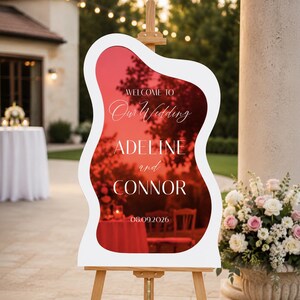 May include: A wavy-edged wedding welcome sign with a reflective red surface, mounted on a wooden easel. The white text reads: WELCOME TO Our Wedding ADELINE and CONNOR 08.09.2026. The sign features a smooth, irregular, organic shape.