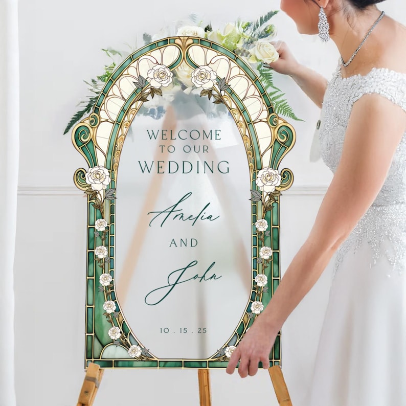 May include: An arched wedding welcome sign featuring a stained glass-style border in shades of green and cream with white rose illustrations. The centre text reads Welcome to our wedding, Amelia and John, 10.15.25, printed on a clear acrylic panel.