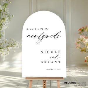 May include: An arched white wedding sign on a wooden easel featuring black text that reads brunch with the newlyweds, NICOLE and BRYANT, and AUGUST 14, 2024. The sign has a smooth surface and a clean, minimalist layout with elegant typography.