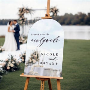 May include: An arched mirror wedding sign on a wooden easel with black text reading: brunch with the newlyweds, NICOLE and BRYANT, AUGUST 14, 2024. The reflective surface displays the surrounding outdoor scenery and wedding decor.