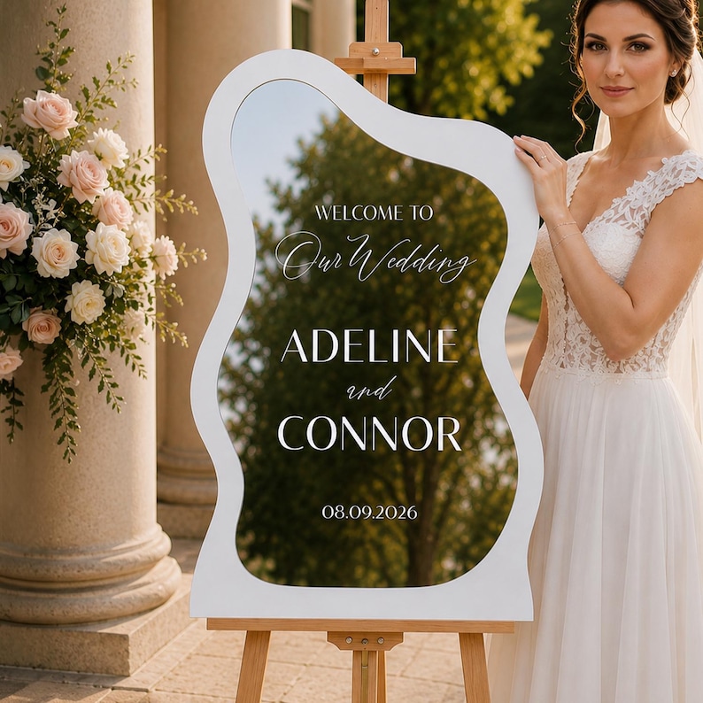 May include: A white, wavy-edged wedding welcome sign on a wooden easel. The sign features the text: WELCOME TO Our Wedding ADELINE and CONNOR 08.09.2026. The surface is reflective, showing a blurred outdoor scene with trees and greenery.